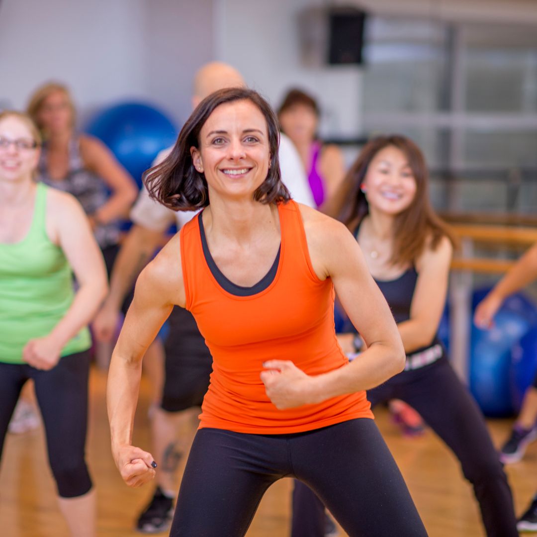 active-visible-leadership-change-management Strong woman wearing orange workout clothes leads an aerobics class. She is determined. The people behind her are following along smiling, slightly out of focus.