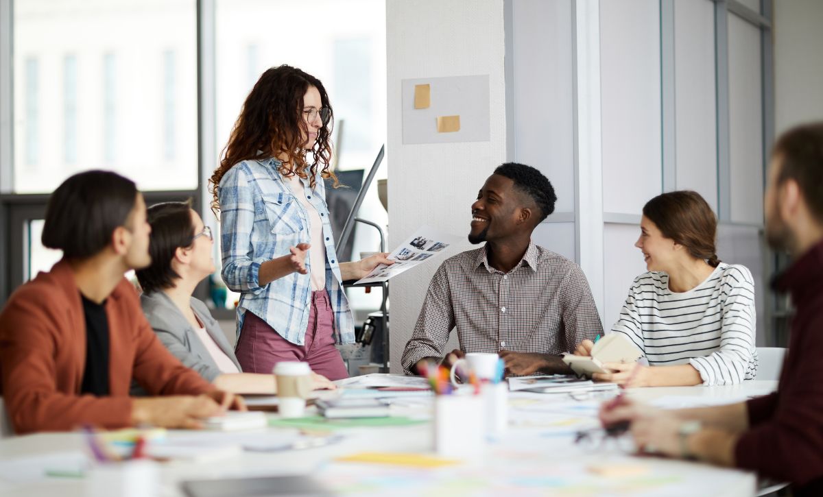 A team of five colleagues gathered around a table. A young woman in casual clothing is presenting an idea while the team listen eagerly.