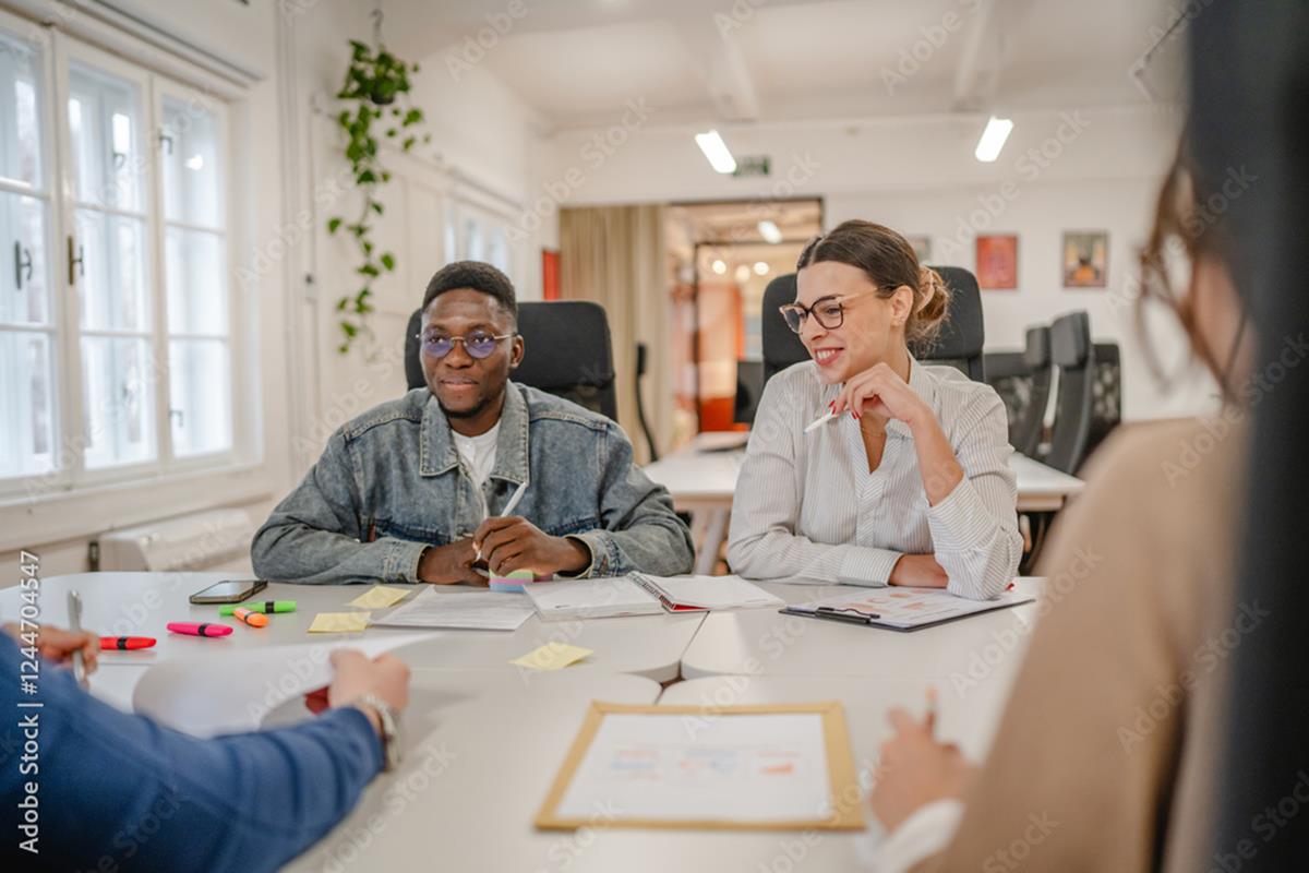 Four people sit around a table in a bright office, discussing documents and using markers. Two people, one in a denim jacket and one in a white shirt, are the main focus, listening and smiling.