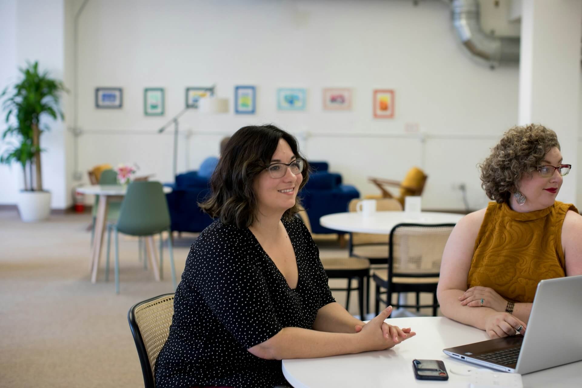 Two women sit at a round table in a modern, open office space. One smiles whilst talking, and the other looks at a laptop. The room features colourful art, plants, and casual seating in the background.