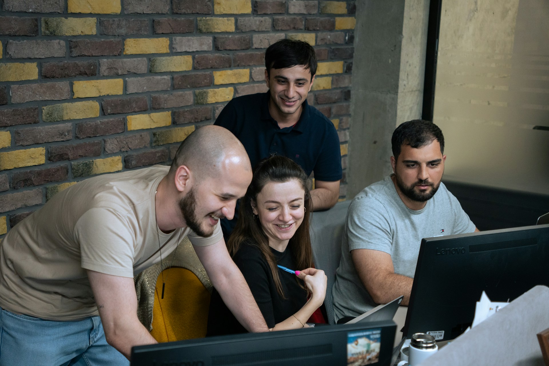 Four people gather around two computer monitors in an office. Three are smiling and looking at one screen whilst one person types, suggesting teamwork and collaboration. Exposed brick wall in the background.