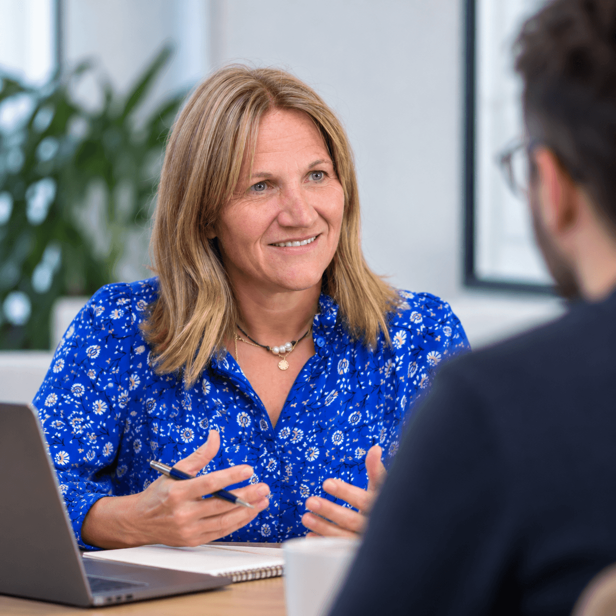 Erica in conversation edit img A smiling woman in a blue patterned blouse speaks with a colleague across a desk, with a laptop and papers nearby in a bright office.