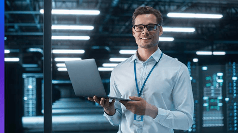 Man wearing glasses and an identification badge holds an open laptop in a modern server room. Server racks and bright overhead lights appear in the background.